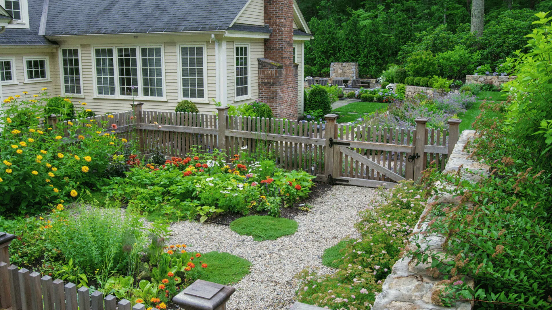 Cottage garden with colorful flower beds, gravel pathways, wooden fencing, and lush greenery surrounding a large house.