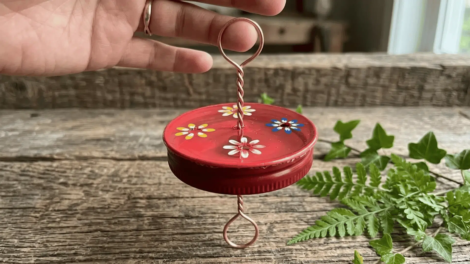 Jar lid DIY hummingbird feeder with painted flowers and copper wire hanger held above a wooden table