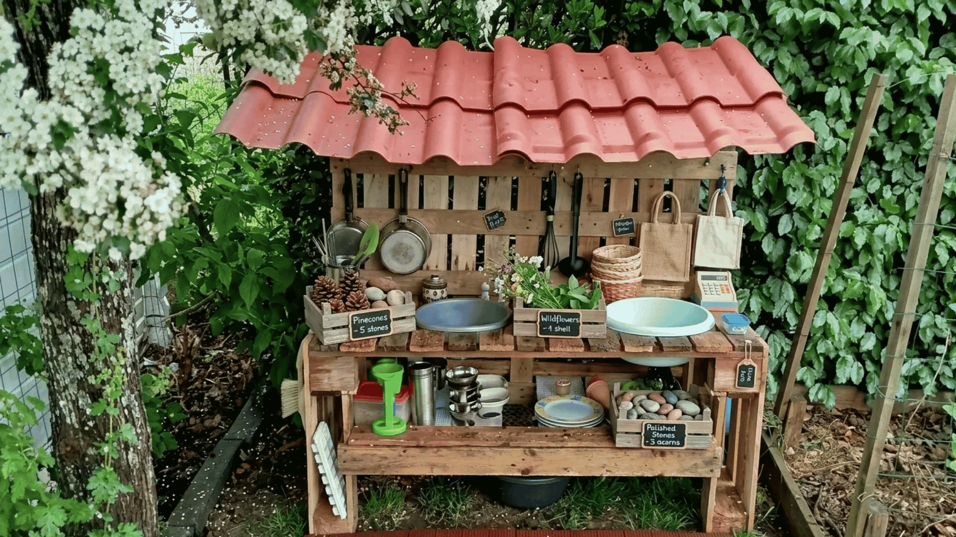 Mini farmers market mud kitchen with nature displays, baskets, labeled produce stands, a sink area, and outdoor role-play shopping setup.