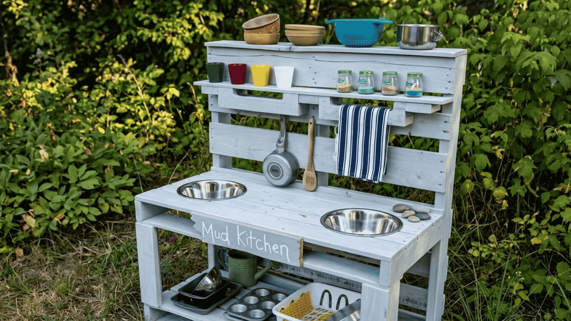 Mud kitchen with two metal sink bowls, hanging utensils, shelves with cups and jars, and storage space underneath in an outdoor garden setting.