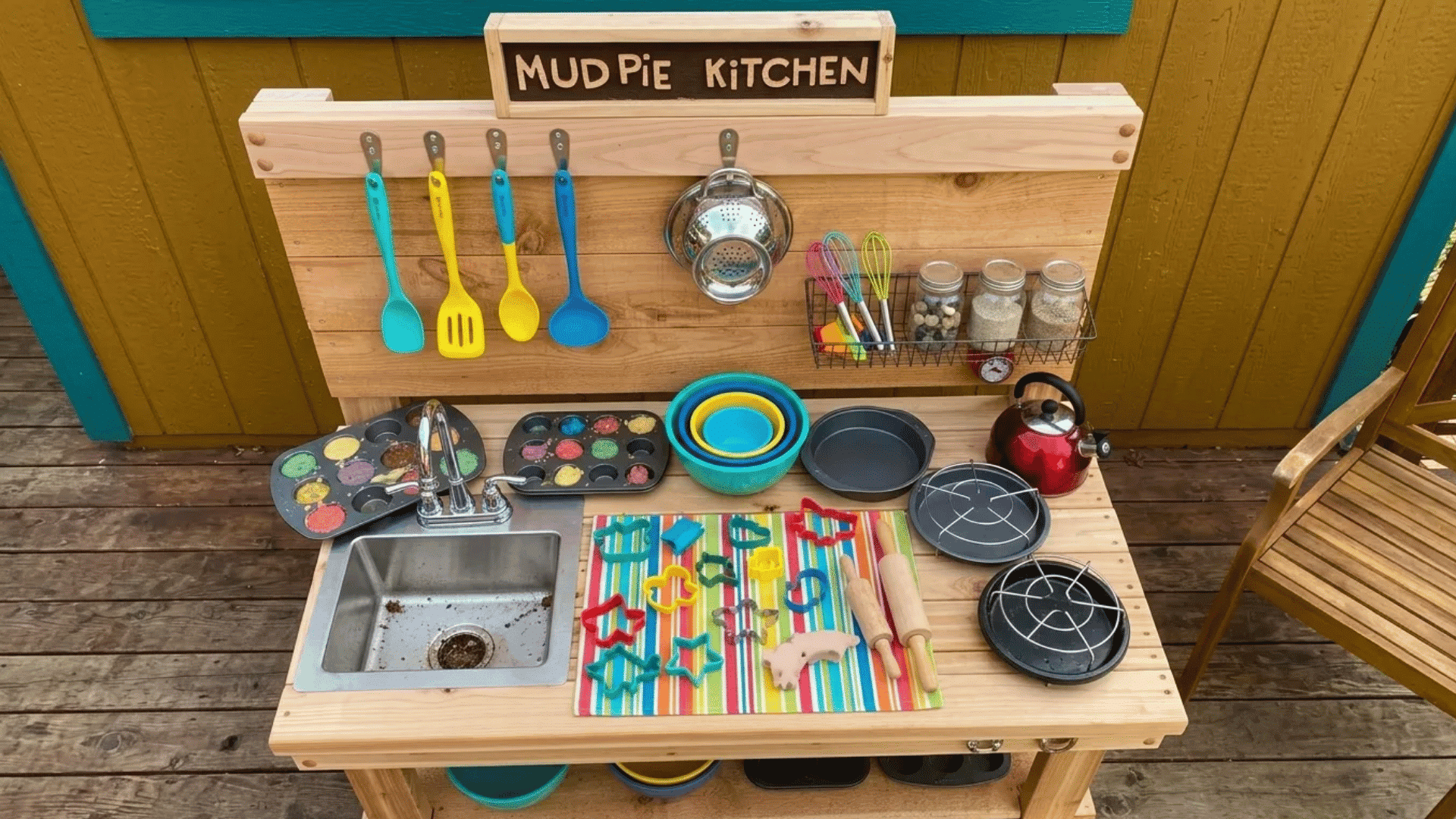 Mud pie bakery mud kitchen with a built-in sink, baking trays, cookie cutters, mixing bowls, hanging utensils, and pretend baking tools for outdoor play.