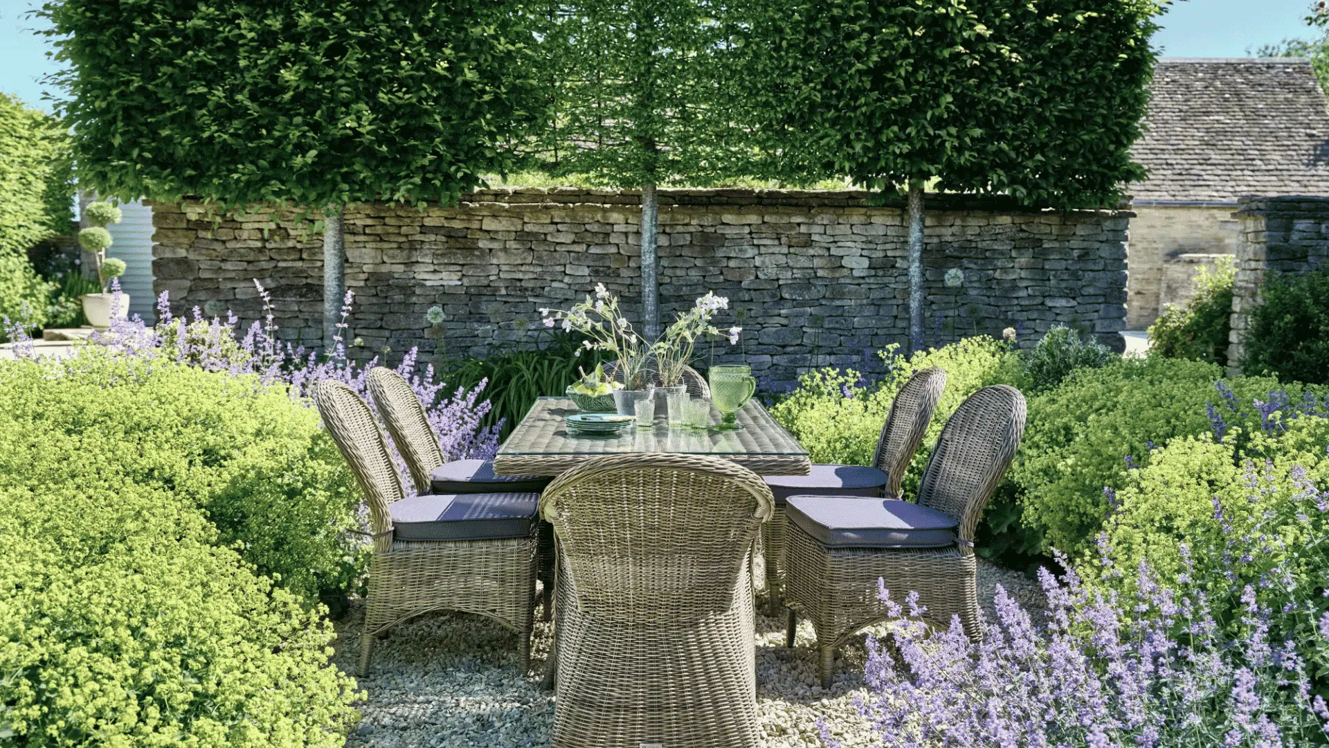Outdoor dining area with wicker chairs and table surrounded by trimmed hedges, lavender flowers, and a stone wall garden.