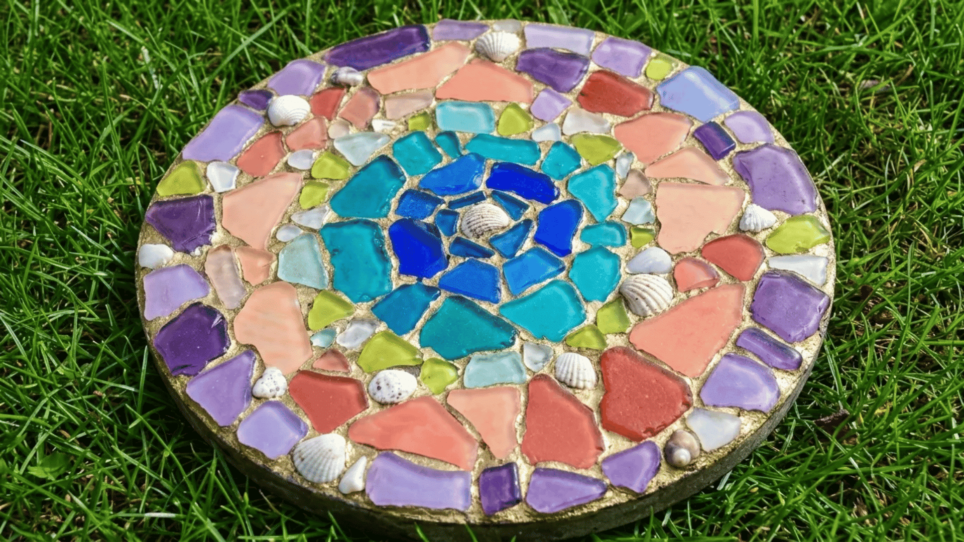 Round sea glass mosaic stepping stone with shells and colorful glass pieces on grass.