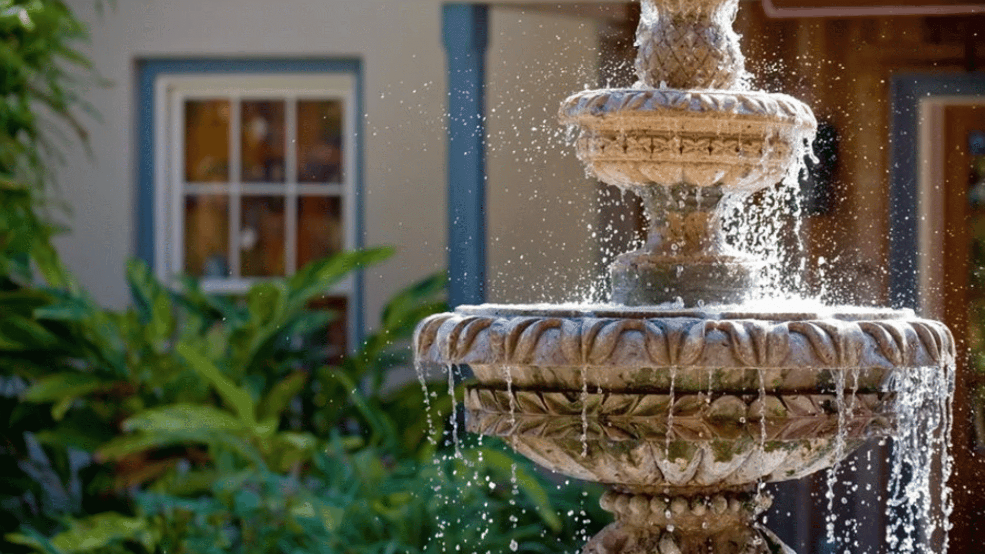 Tiered stone fountain with flowing water in a garden courtyard surrounded by green plants and a blurred house background.
