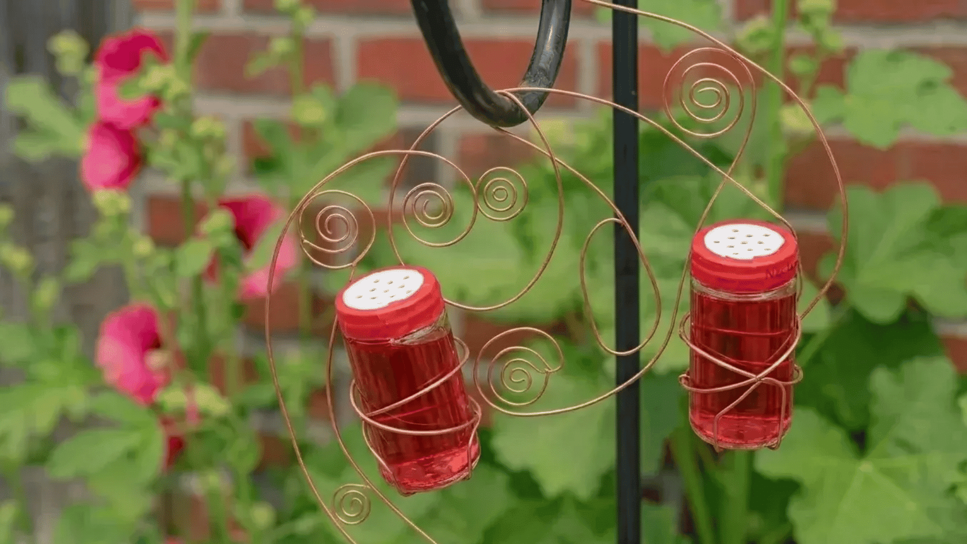 Two small spice jar diy hummingbird feeders with red lids hanging from copper wire in a garden