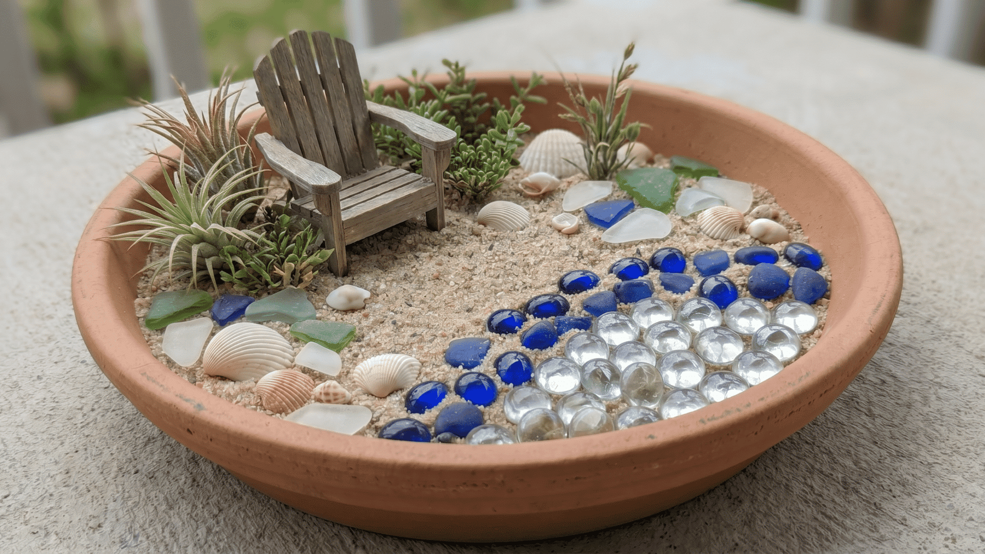 Mini beach fairy garden in a shallow bowl with sand, shells, blue stones, a tiny chair, and small plants