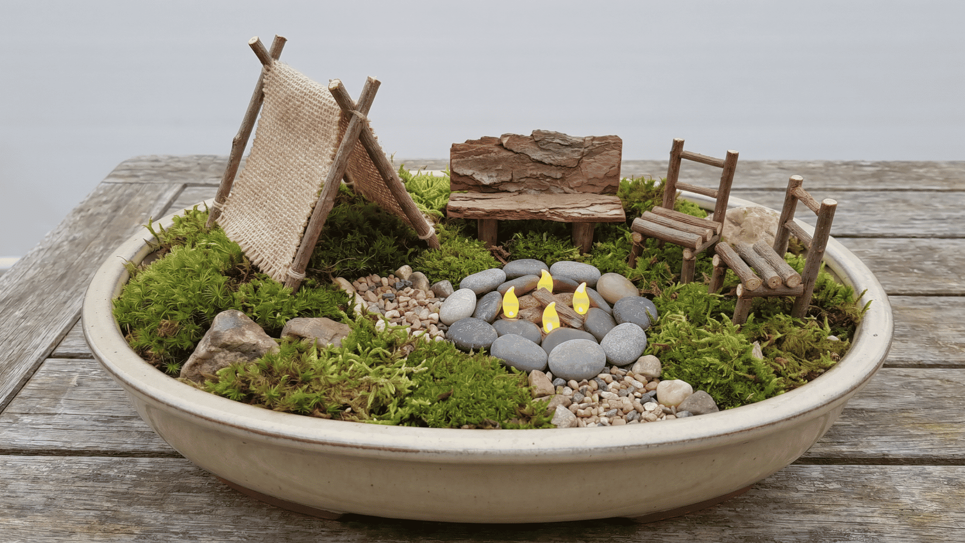 Small camping fairy garden with a twig tent, pebble fire ring, bark bench, chairs, and moss ground