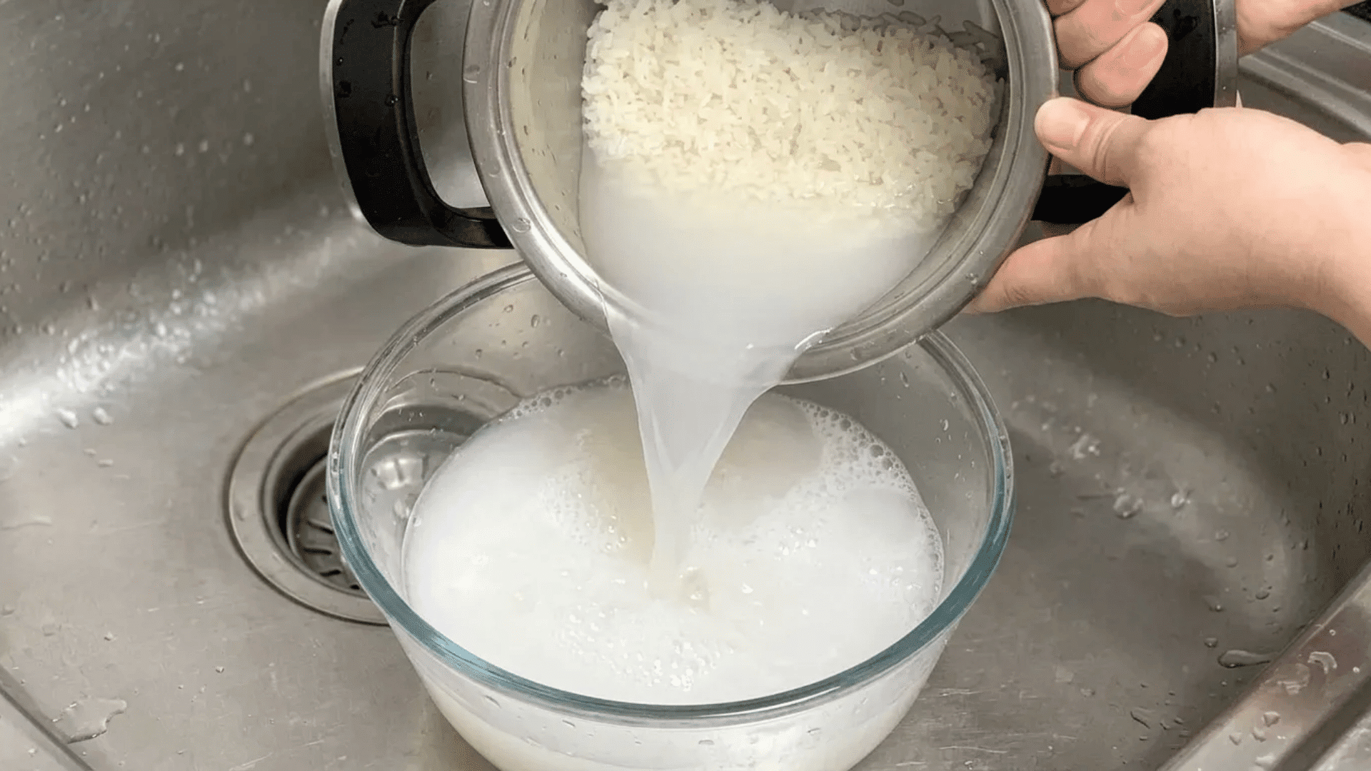 Cloudy rice wash water being poured from a pot into a glass bowl beside rinsed white rice in a sink