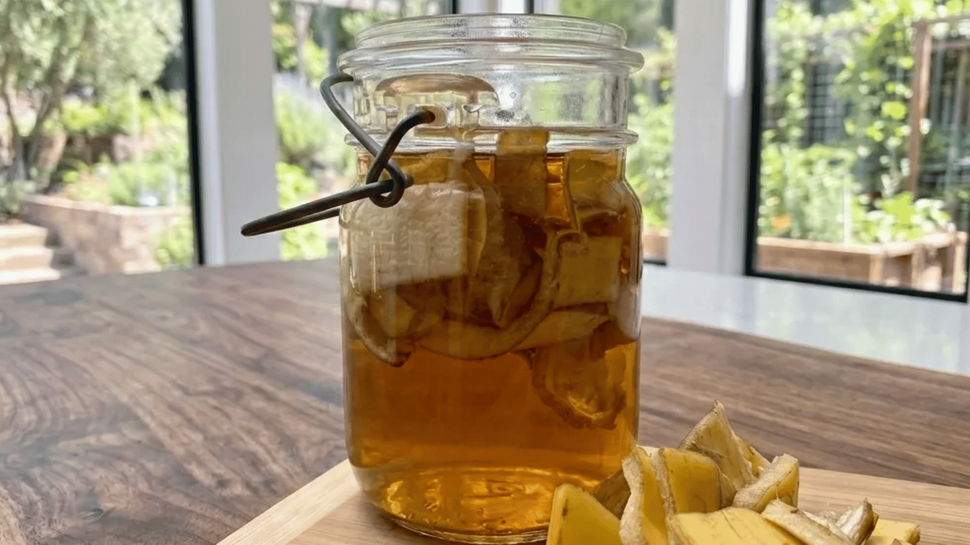 Glass jar filled with banana peel water on a wooden table beside fresh banana peels near a garden window