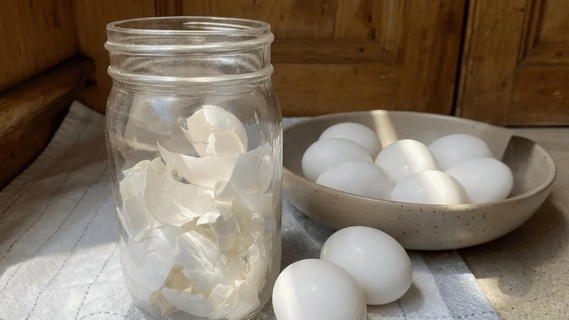 Glass jar filled with crushed eggshells beside whole eggs on a kitchen towel and bowl